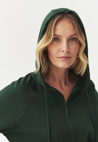 Sudadera verde con cremallera y capucha ajustable, fabricada en un tejido suave y con mangas raglán. Diseño simple que resalta un estilo casual.