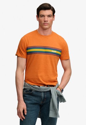 Mand iført orange t-shirt med horisontale grønne, gule og blå striber, blå jeans og en ternet skjorte bundet om livet.