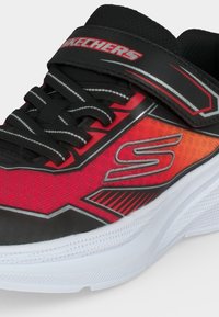 Μαύρο και κόκκινο αθλητικό παπούτσι Skechers με λευκή σόλα, μαύρα κορδόνια, ιμάντα με βέλκρο και λογότυπο στην πλευρά και στον ιμάντα.