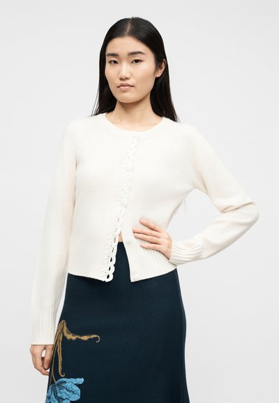 Νεαρή γυναίκα με μακριά σκούρα μαλλιά, φορώντας μια λευκή ζακέτα με κουμπιά και μια σκούρα φούστα με μπλε λουλουδένιο σχέδιο, στέκεται μπροστά σε ένα απλό φόντο.