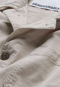 Veste en coton beige avec fermeture à boutons, une poche et des détails de coutures apparentes. Étiquette indiquant "ARMEDANGELS." Texture douce.