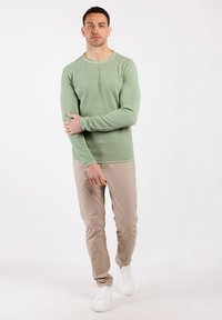 Uomo vestito con una camicia a maniche lunghe verde chiaro, pantaloni beige e sneakers bianche, in piedi contro uno sfondo bianco uniforme con il braccio sinistro che tiene il braccio destro.