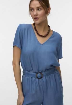 Vrouw draagt een blauwe korte mouw jumpsuit met V-hals, een ronde riemgesp en een donkere kralenhalsketting, kijkend naar links.