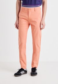 Peach-colored pants made of durable fabric, featuring a straight fit, standard waistband, and front pockets, paired with black athletic shoes.