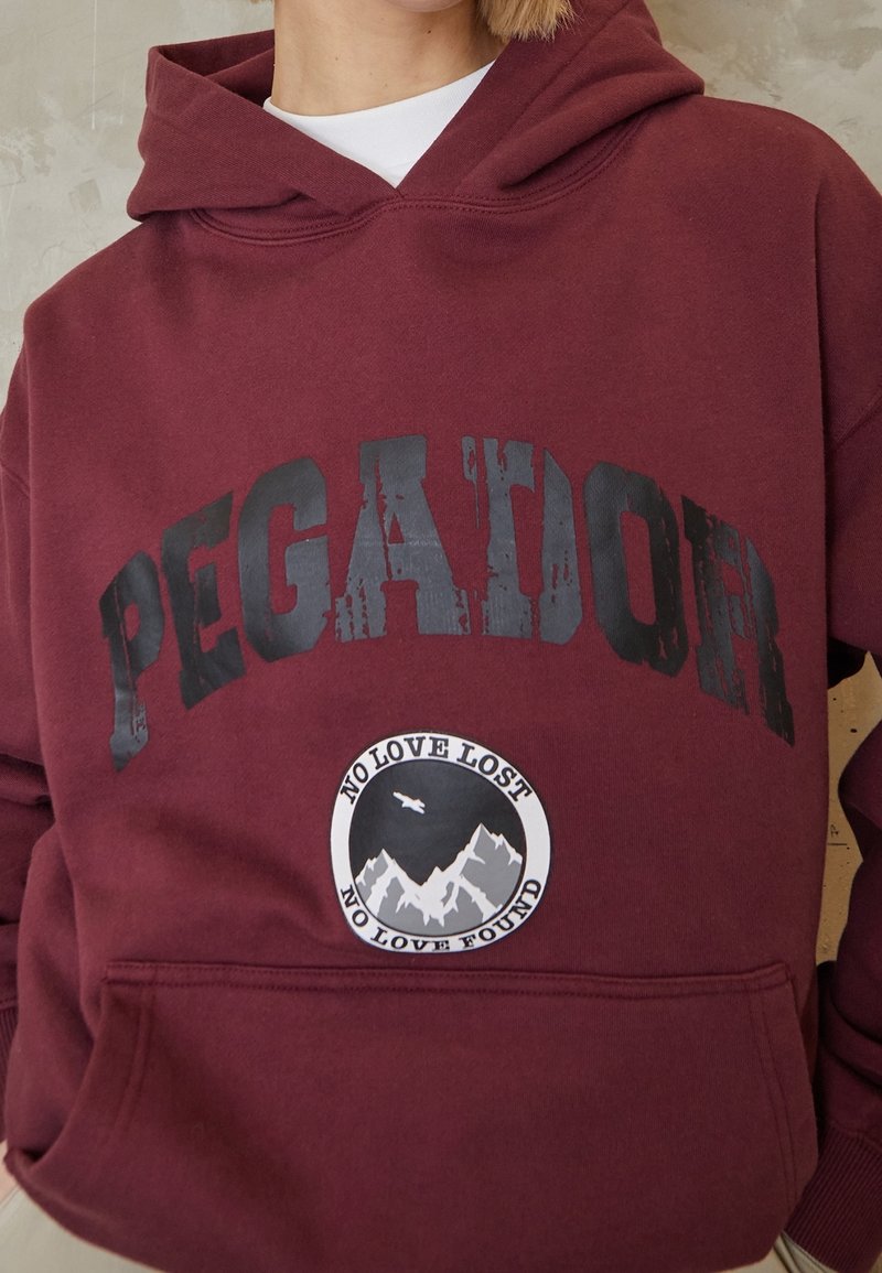 Osoba v bordovém mikině s kapucí s nápisem "PEGATOR" v černých písmenech a kulatou nášivkou s horami, ptákem a textem "NO LOVE LOST NO LOVE FOUND."