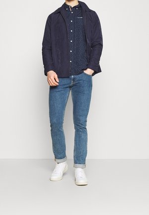 Navy lightweight jacket over a navy polka dot shirt, paired with slim blue jeans and white sneakers, displaying a casual outfit.