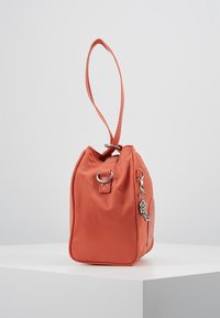 Mała pomarańczowa torebka na ramię z paskiem na nadgarstek, srebrnymi metalowymi pierścieniami i wiszącym srebrnym charmsu, wyeksponowana na białym piedestale na tle jednolitym.