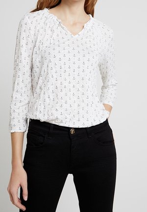 Kvinde iført en hvid bluse med lange ærmer med små sorte ankerprint og sorte bukser, den ene hånd i lommen.