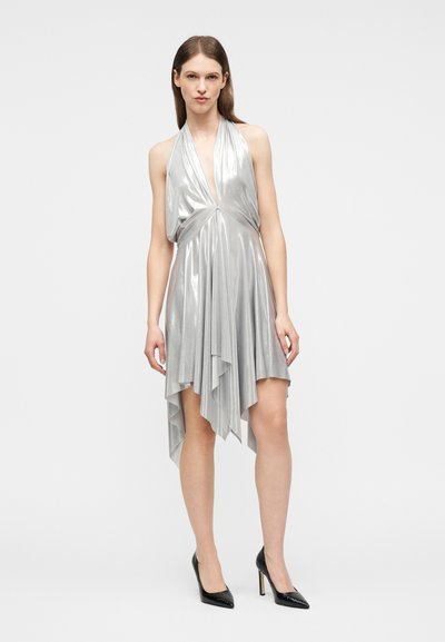 Γυναίκα που φοράει αμάνικο ασημί μεταλλικό φόρεμα με έναν ώμο και ασύμμετρο τελείωμα, μαζί με μαύρα παπούτσια με ψηλά τακούνια, που στέκεται μπροστά σε ένα απλό φόντο.