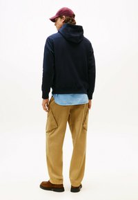 Marineblauer Hoodie mit Kängurutasche, hellblaues Hemd darunter, beigefarbene Cargo-Hose und kastanienfarbene Mütze. Braune Schuhe mit dunklen Sohlen.