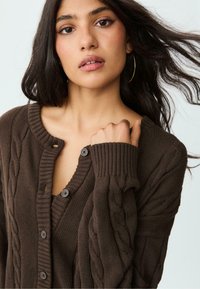 Brauner Strickcardigan mit einem Zopfmuster, gerippten Bündchen und Knopfverschluss. Hat einen V-Ausschnitt und strukturierte Details.