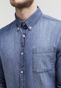 Camisa de denim en azul oscuro. Presenta un cuello abotonado, siete botones y un bolsillo en el pecho. Tela texturizada con un acabado suave.