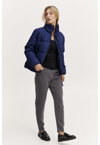 Marinblå puffjacka med hög krage, quiltat mönster och dragkedjestängning, kombinerad med grå byxor och svarta loafers.