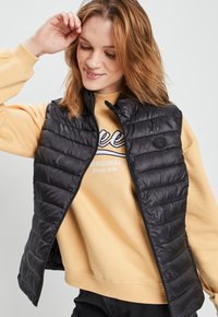 Zwarte gewatteerde vest met een hoge kraag en rits; gedragen over een gele sweatshirt met een grafische print. Gladde, glanzende textuur.