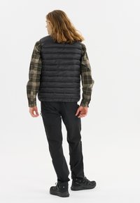 Schwarze Steppweste mit einem gesteppten Design, getragen über einem grün karierten Hemd; kombiniert mit schwarzen Hosen und robustem schwarzem Schuhwerk.