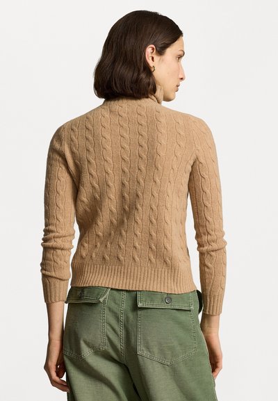 Bež kabeli kudumitega sviiterit kõrge kaelusega, sooniliste äärte ja slim-fit varrukatega, koos roheliste pükstega, millel on tagataskud.