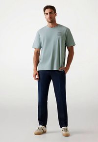 Svijetlozelena pamučna majica kratkih rukava s okruglim izrezom i malim grafikonom na lijevoj strani prsa, u kombinaciji s tamnoplavim trapericama i bež tenisicama.