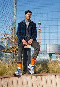 Graue und weiße Turnschuhe mit blauen Schnürsenkeln, olivgrüne Cordhosen, ein grauer Rollkragenpullover und ein blau kariertem Mantel, kombiniert mit orangefarbenen Socken.