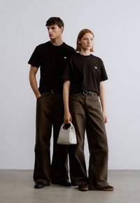 Μαύρα βαμβακερά μπλουζάκια, φαρδιά καφέ παντελόνια και μια μικρή λευκή τσάντα χειρός. Απλός σχεδιασμός χωρίς σχέδια, σε ουδέτερο φόντο.