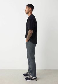 Camiseta negra de manga corta, pantalones gris rectos y zapatillas negras con detalles grises; perfil de pie contra un fondo blanco.