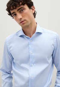 Giovane uomo con capelli scuri e ricci che indossa una camicia button-up a righe sottili di blu chiaro su uno sfondo chiaro e semplice.