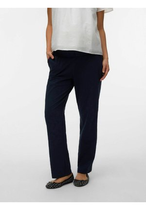 Moteris dėvi tamsiai mėlynas tiesias kelnes, juodus ir baltus tekstūruotus baleto batelius bei baltą trumpomis rankovėmis palaidinę su subtiliu gėlių siuvinėjimu.