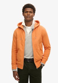 Orange zip-up hoodie med to frontlommer, lagd oven på en lys grå polo. Modellen bærer mørkegrønne bukser og viser et ur på venstre håndled.