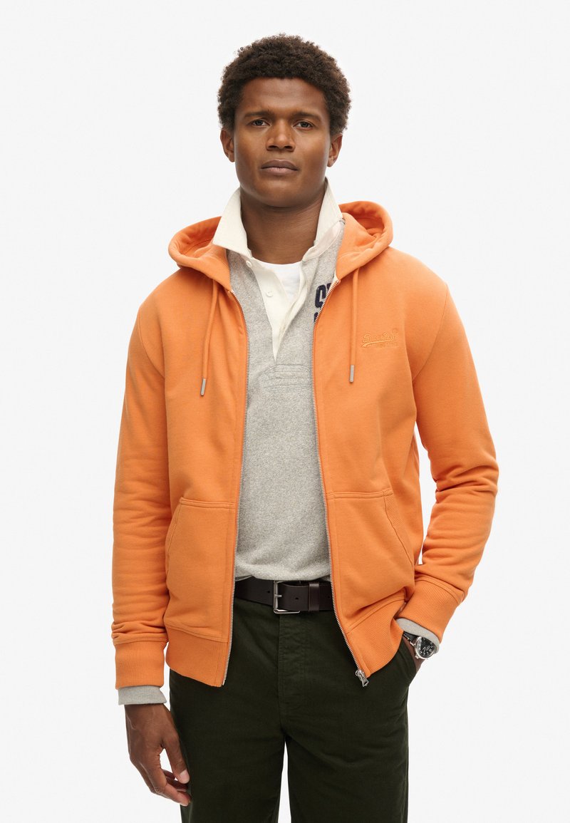 Orange zip-up hoodie med to frontlommer, lagd oven på en lys grå polo. Modellen bærer mørkegrønne bukser og viser et ur på venstre håndled.