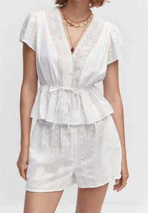 Woman wearing a white eyelet lace short-sleeve blouse with tie waist and matching high-waisted shorts, accessorized with layered necklaces.