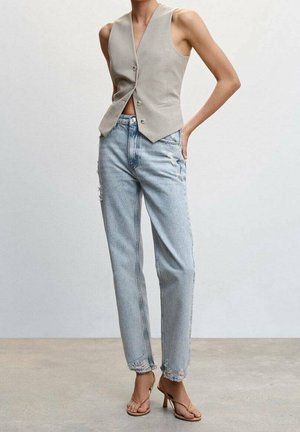 Nainen, jolla on yllään hihaton beige liivi, vaaleat kuluneet korkeavyötäröiset farkut ja vaaleanruskeat korkokengät, seisoo harmaalla lattialla yksinkertaista seinää vasten.