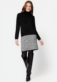 Schwarzer Rollkragenpullover mit langen Ärmeln, kombiniert mit einem schwarz-weiß gemusterten Rock. Glänzende schwarze kniehohe Stiefel vervollständigen das Outfit.