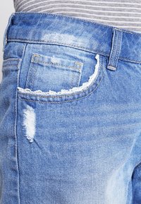 Primer plano de unos jeans de mezclilla azul claro con un pequeño detalle desgastado y un bolsillo delantero con borde de encaje, combinados con una blusa gris a rayas.