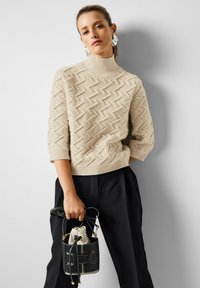 Frau in einem beigen strukturierten Rollkragenpullover und schwarzen Hosen, die eine kleine schwarze Bucket-Bag mit weißer Naht hält.