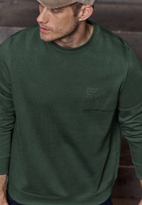 Dunkelgrüner Kapuzenpullover aus strukturiertem Stoff, mit einem Rundhalsausschnitt und dezentem Druck-Logo auf der Brust. Entworfen für Wärme.