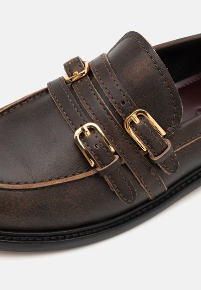 Mocasín de cuero marrón oscuro con acentos de doble hebilla dorada, una textura suave y costuras en contraste a lo largo de los bordes y la suela.