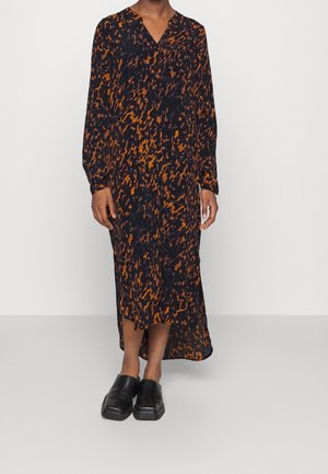 Osoba nosi czarną i pomarańczową sukienkę midi z abstrakcyjnym wzorem, z długimi rękawami i dekoltem w kształcie litery V oraz czarne wsuwane buty z kwadratowym noskiem.