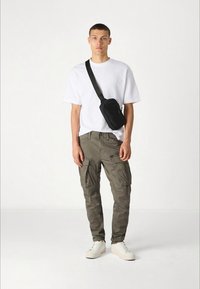 Witte katoenen T-shirt, olijfgroene cargo broek met zakken, zwarte schoudertas en witte sneakers. Eenvoudige, casual outfit.