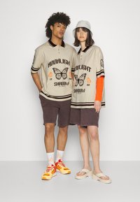 Beige oversized polo skjorte med sorte striper, sommerfugltrykk og oransje detaljer. Modellen kombinerer den med mørke shorts og fargerike joggesko.