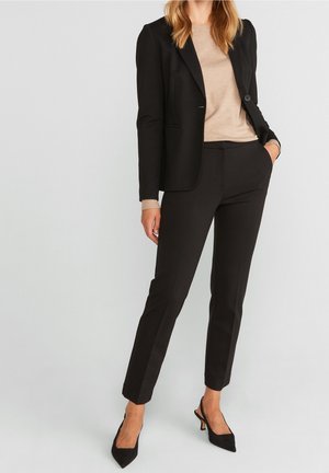 Femme portant un blazer noir cintré, un pantalon slim assorti, un haut beige à manches longues et des talons noirs à bout pointu, debout avec une main dans la poche.