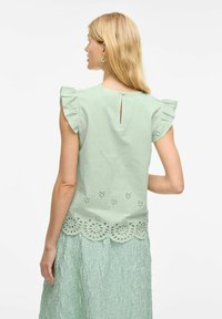 Svetle zelený top s krátkým rukávem a volánkovými detaily, který má otvor na zádech a laserem vyřezávaný květinový vzor podél lemu.