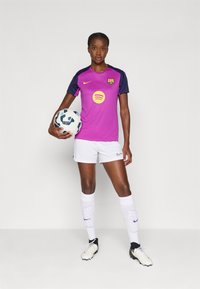 Lilla og marineblå fotballdrakt med korte ermer, hvite shorts og sokker. Spilleren holder en fotball med svarte og blå detaljer.