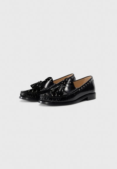 Mocassins en cuir verni noir avec une frange texturée, un détail de nœud et des clous métalliques le long du col. Semelle plate, design à bout rond.