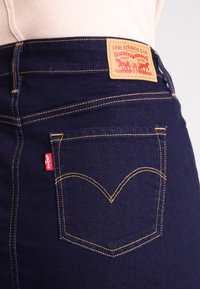 Mörkblå denimjeans med gula sömmar, med en läderpatch på ryggen och en röd logotagg på sidofickan.