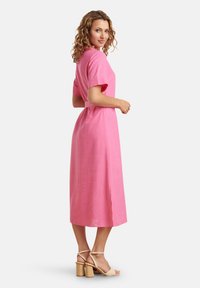 Rosa Leinenkleid mit kurzen Ärmeln, tailliertem Schnitt und seitlichen Schlitzern. Das Modell trägt beigefarbene Absatzsandalen und zeigt ein mittellanges Design.