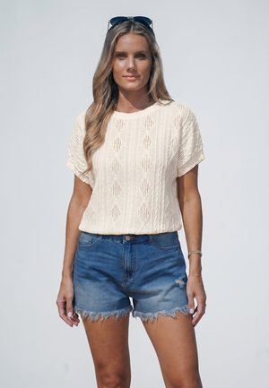 Vrouw met lang haar met zonnebril op haar hoofd, crème gebreide korte mouw top en versleten denim shorts, staand tegen een effen achtergrond.