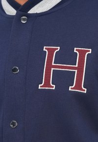 Tamno plavi dres sivi rebrasti ovratnik, sadrži veliki crveni logo "H" s teksturiranom vezom i akcentom srebrnog dugmeta na kopčanju.