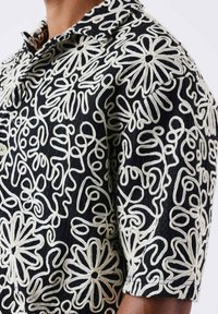 Camisa negra de manga corta con intrincados bordados florales y abstractos en blanco. Tejido texturizado con cuello y detalles de botones.