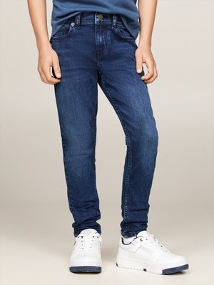Jeans slim-fit blu scuro con leggere scoloriture, cinque tasche e passanti per cintura, abbinati a sneakers bianche con accenti blu navy.