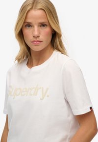 Hvid bomulds t-shirt med et forhøjet beige "Superdry" logo, rund hals, korte ærmer og et rødt og sort mærke på venstre ærme.