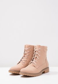 Ett par beige snörstövlar med präglade blommönster och tjocka gummisolen, mot en enkel vit bakgrund.